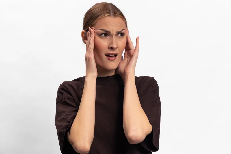 Young woman with brown hair in dark shirt shows confused expression touching temples. Isolated on white background. Concept of doubt, uncertainty, and indecision in everyday life.