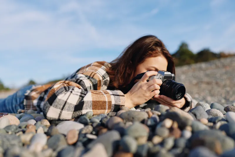 Female photographer lying on a rocky beach, wearing a plaid shirt, focusing through a handheld camera to capture coastal landscapes under a clear blue sky and nearby pebbles.