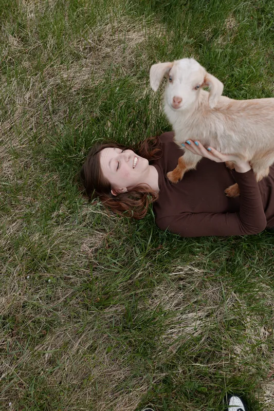 Casual lifestyle scene shows a woman lying on grass outdoors, a young goat kid perched on her chest, sharing an affectionate moment amid meadow, showcasing simple joy and bond with animals in a