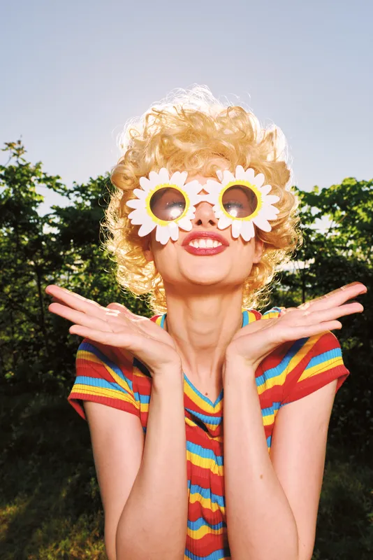 A woman in retro colorful dress stands outdoors with flower sunglasses, showcasing lifestyle, portrait, and playful mood in a sunlit park. Bright, cheerful, and fashion-forward, the scene evokes