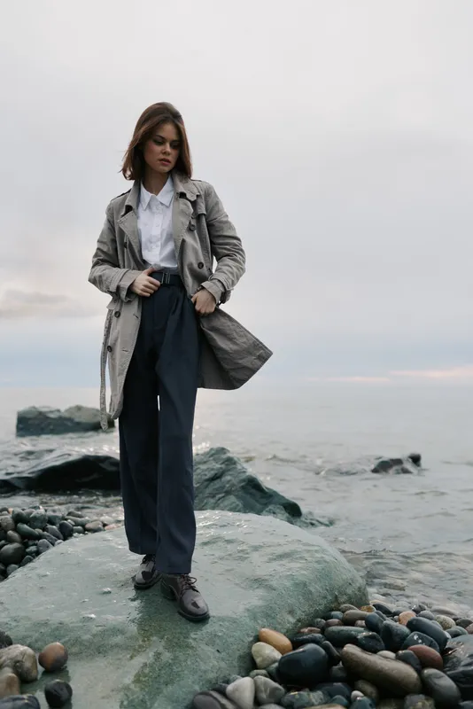 Stylish woman in long coat and trousers stands on rocky shore near calm sea under overcast sky. Fashion portrait with natural colors and serene coastal background concept.