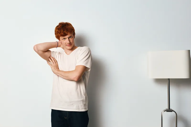 Young man with red hair wearing white t shirt and dark pants stretching arm behind head standing near lamp indoors on white wall background in casual style portrait.