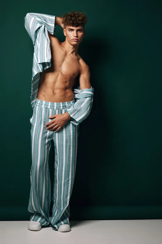 Young man posing in striped pajamas with open shirt showing toned abs, standing against dark green background in studio with confident expression and casual posture.