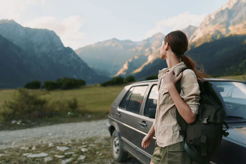 Young woman with backpack standing near car looking at mountain landscape, outdoor adventure, casual outfit, summer travel, nature exploration
