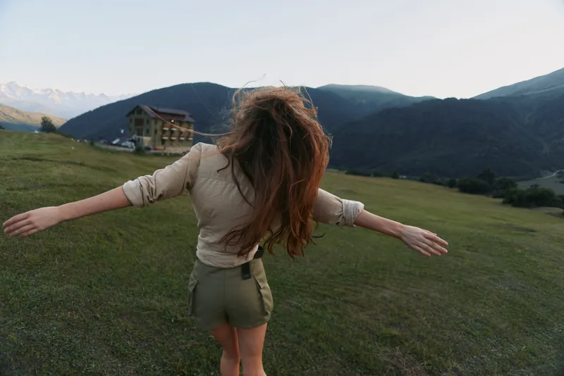 Young woman with long red hair enjoys freedom with outstretched arms in a green meadow surrounded by mountains during a clear day.