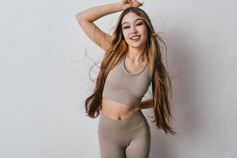 Young woman in beige workout clothes smiling and posing with long hair on white background. Fitness, exercise, active lifestyle and healthy youth with happiness and energy.