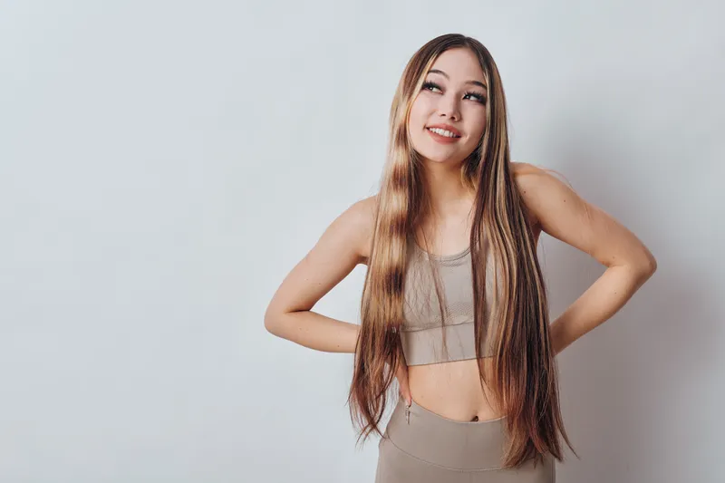 Young woman with long straight hair wearing casual tank top and sweatpants stands with hands on waist smiling against light background.