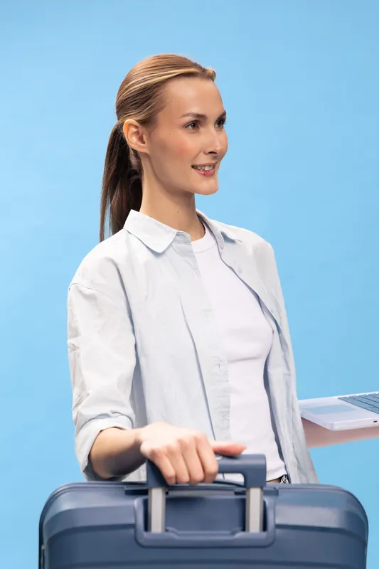Young woman with ponytail and casual white shirt holds blue suitcase handle and laptop, smiling. Travel, business trip, and technology concept on blue background with optimistic mood.
