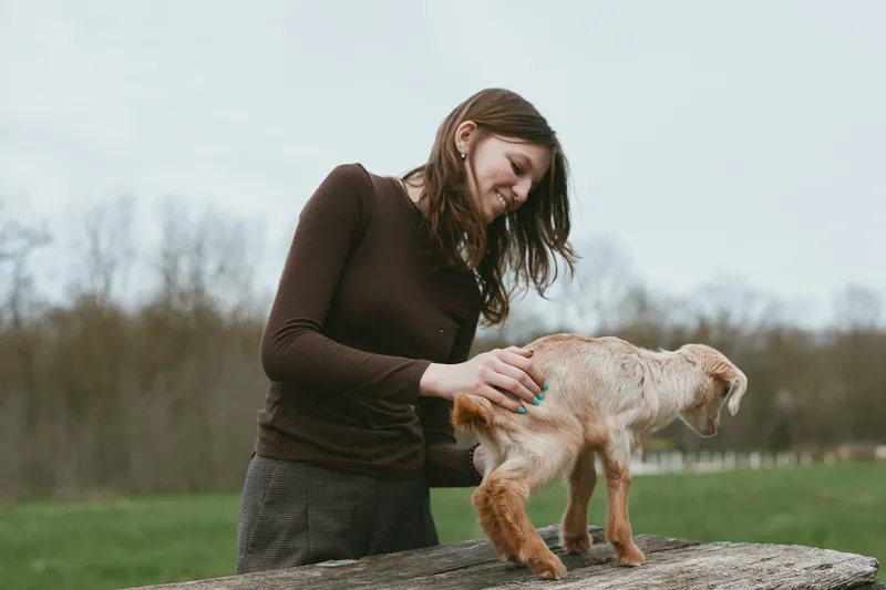 Young woman smiles softly while guiding a goat kid over a wooden barrier in an outdoor setting, capturing a warm lifestyle moment of care, connection, and animal companionship under natural light.