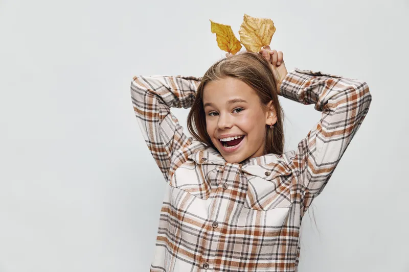 Girl in plaid shirt smiling and holding autumn leaves behind head in playful pose with white background. Young person enjoying fall season with happy expression and natural light.