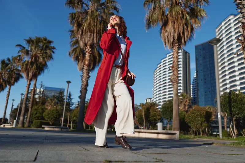 Woman wearing a red coat and white pants walks through a sunny urban city plaza with palm trees and modern architecture, capturing confident fashion and street style.