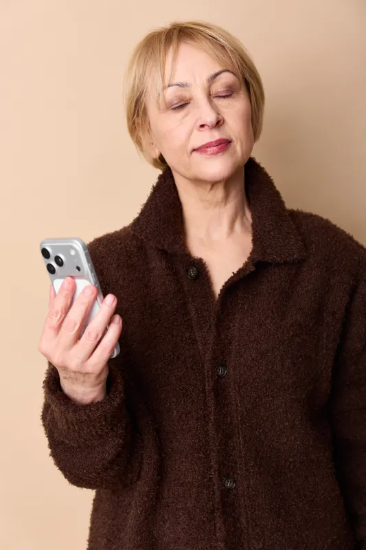 Woman senior portrait holding smartphone in hand, eyes closed, beige studio background, dark coat, concept technology communication, closeup calm moment, indoor wellness