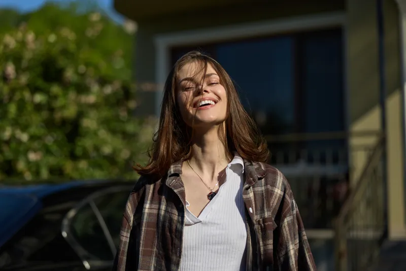 Young woman smiles in sunlight outdoors wearing a plaid shirt, relaxed casual vibe, confident pose, warm breeze, natural colors, relatable lifestyle moment