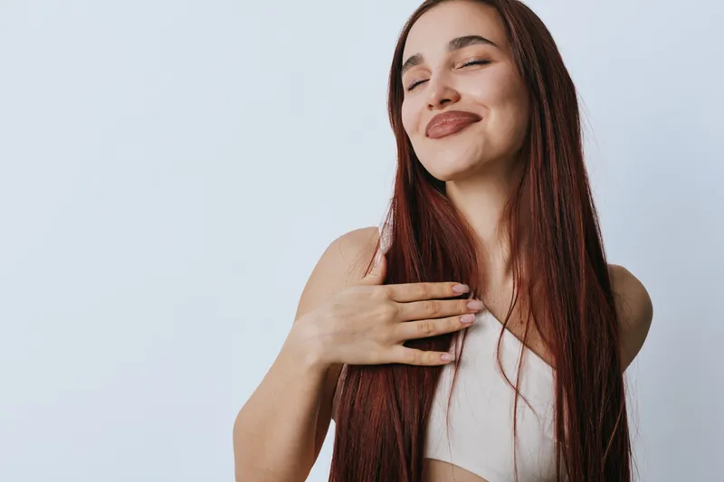 Woman with long hair smiling and holding hand on chest with closed eyes for gratitude and self appreciation on light background. Positive emotion and calm expression.