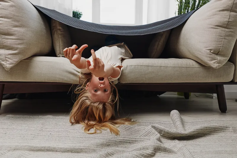 Playful child hides under couch blanket with upside down head and outstretched arms on carpet in cozy living room. Concept for childhood fun, imagination, and home playtime.