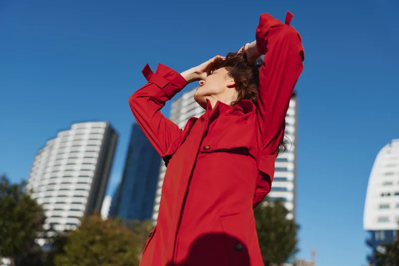 Woman in a bright red coat stands outdoors, looking up at a clear blue sky, with modern city buildings in the background. Fashionable urban style and confident mood.
