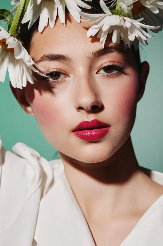 Portrait of stylish young woman with daisy flower crown and vibrant pink lipstick. Fresh makeup highlights smooth skin in high contrast studio lighting. Beauty fashion creative concept.