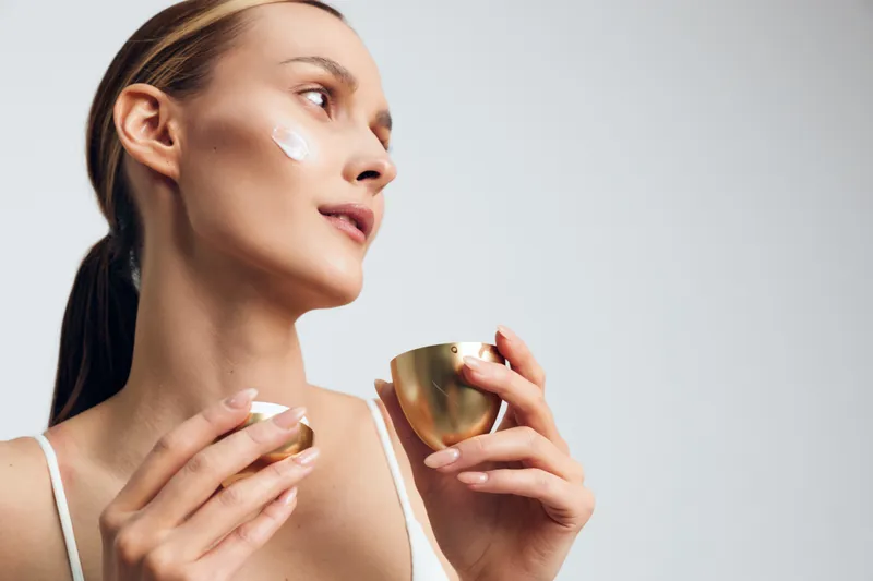 Woman face skincare cream beauty self care hydration spa concept, moisturizer on cheeks, holding cups with water, calm profile expression, bright minimal studio background