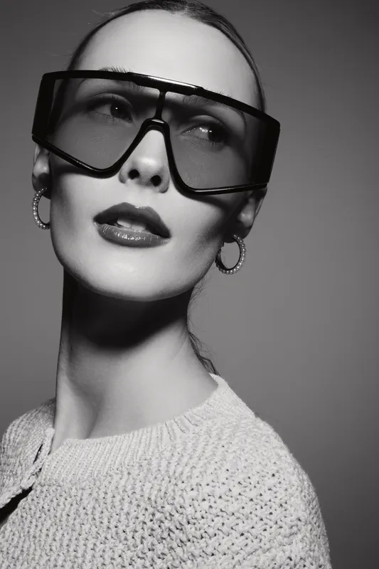 Elegant white woman with pale skin wearing oversized sunglasses and hoop earrings poses in studio. Fashion portrait with stylish accessories and textured sweater concept.