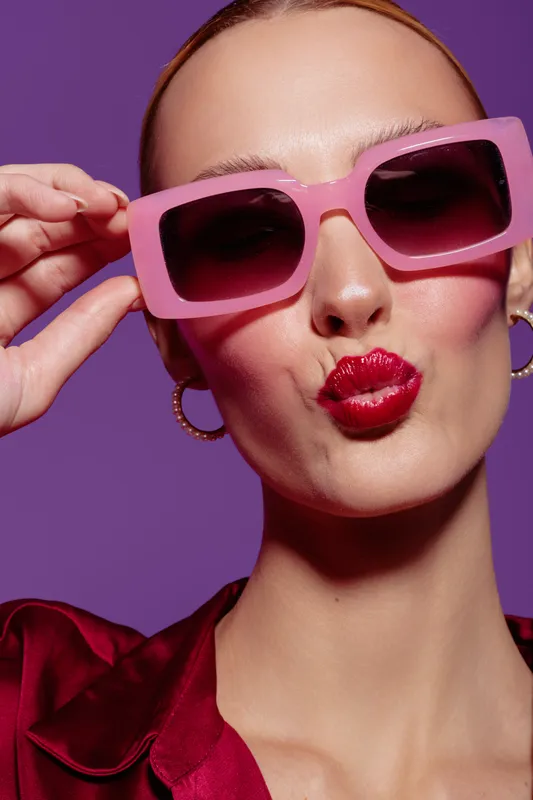Close-up of a fashionable woman wearing oversized pink sunglasses and red lipstick making a playful kiss face. Purple background and satin red blouse create a vibrant, stylish mood.