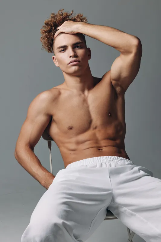 Young muscular man with curly hair posing shirtless in white pants sitting on chair with hand on head against gray background in studio lighting.