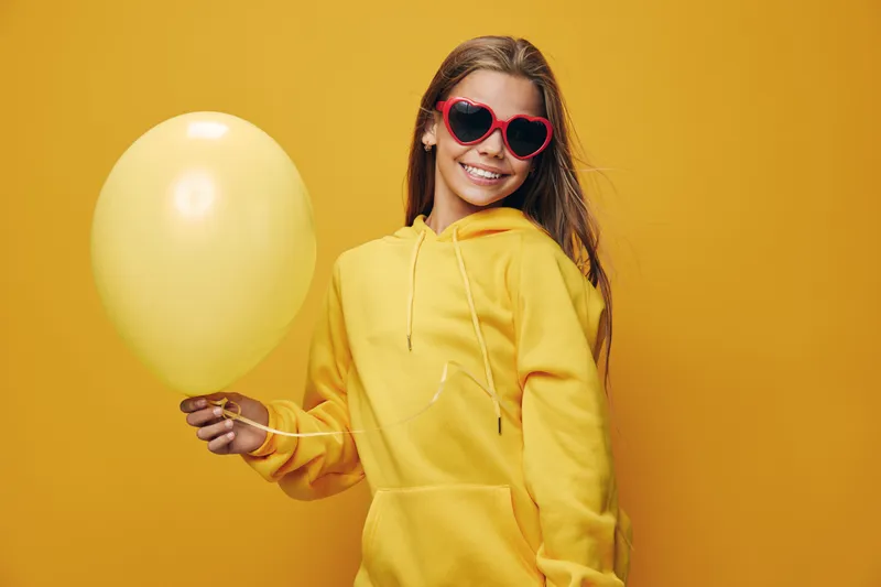 Girl wearing yellow hoodie and heart shaped sunglasses holding yellow balloon on yellow background with smile for happiness and celebration.