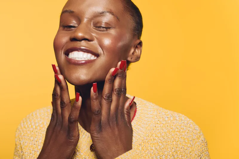 Joyful black woman with dark skin and red nails smiles with closed eyes, touching her face on yellow background. Concept of happiness, beauty, self-care, and positive emotions.