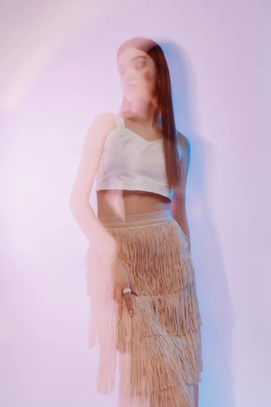 Young woman in a white crop top and fringed skirt poses with soft lighting and motion blur. Concept of beauty, fashion, and modern style in a studio setting.
