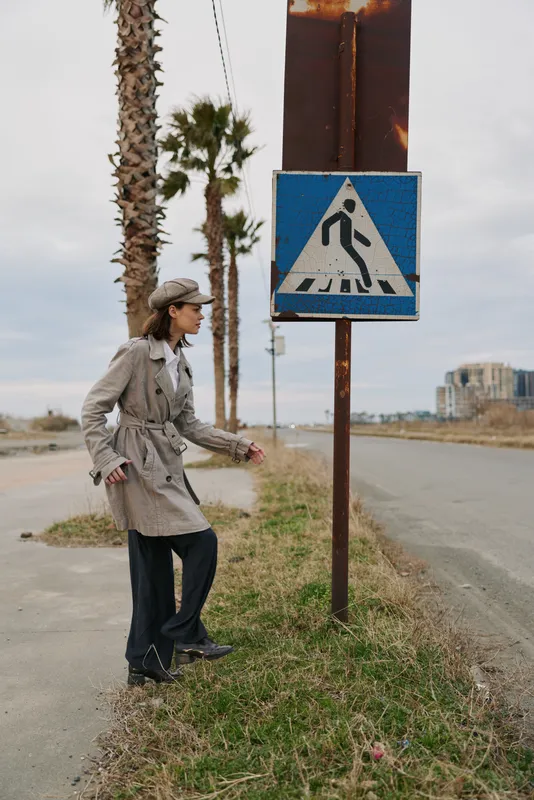 A young woman in a trench coat and hat stands near a pedestrian crossing sign on an empty road with palm trees in the background. Urban outdoor lifestyle concept.