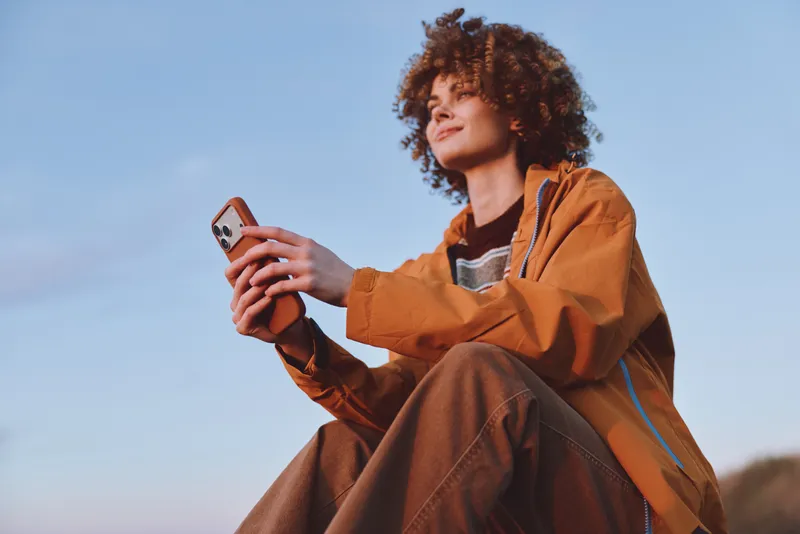 Woman lifestyle outdoors, curly haired woman in brown jacket holds smartphone, looks into distance, relaxed pose, sunny sky, soft breeze feeling, warm natural colors, open field background
