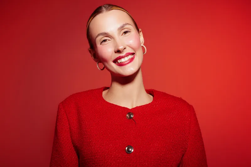 Smiling woman with slicked back hair wearing red sweater poses against red background. Bright makeup and hoop earrings create stylish and confident portrait with warm lighting.