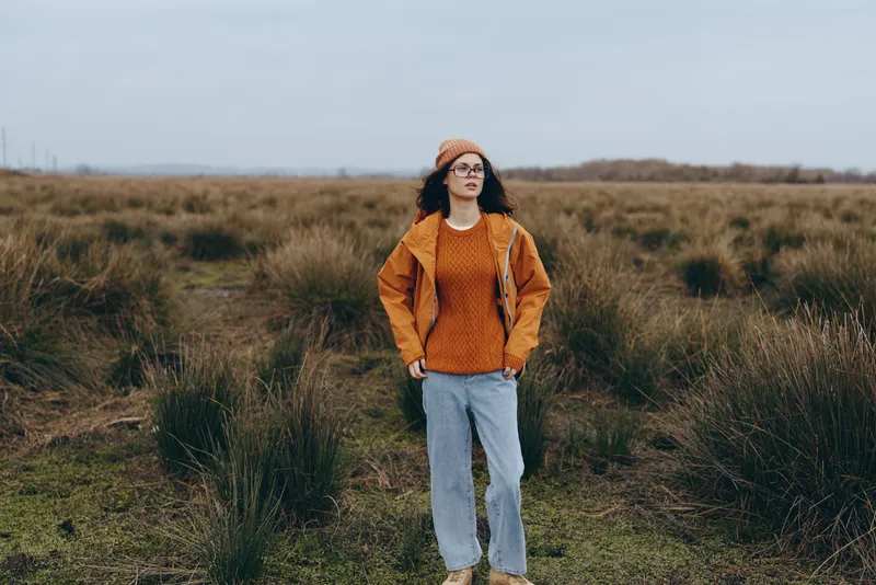 Woman lifestyle portrait outdoors in a grassy field, wearing orange sweater, knit beanie and sunglasses, hands in pockets, standing confidently with calm expression in daylight rural