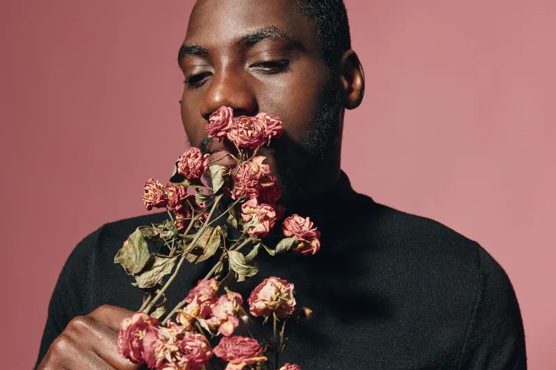 African man with closed eyes holding dried pink roses in hands against pink background in creative conceptual visual style. Portrait of thoughtful black male with flowers in studio.