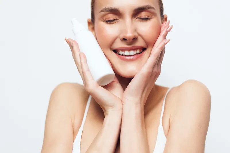 Smiling woman with clear skin holding white cosmetic bottle near face, closed eyes, soft natural light, minimal makeup, skincare routine, fresh beauty concept.