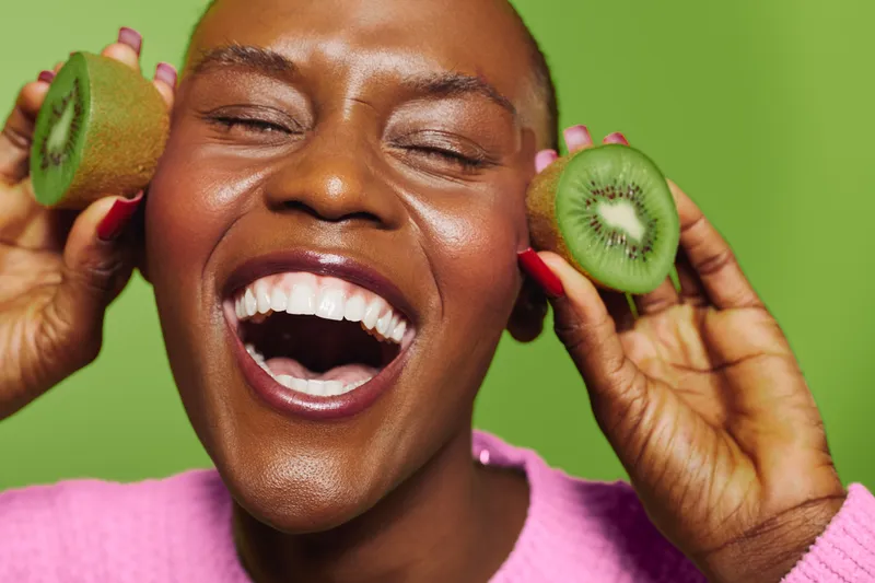 Happy African woman with dark skin laughing and holding two halves of a kiwi fruit near her face. Bright green background and pink sweater create lively and fresh mood for healthy lifestyle concept.