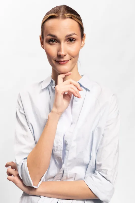Confident woman in white shirt poses with hand on chin against white background. Portrait of thoughtful female model with natural makeup and sleek hair. Concept of business and success.