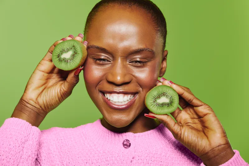 Smiling dark skinned African woman holds two halves of fresh kiwi near face, wearing pink sweater. Green background, healthy eating, natural beauty and fruit concept with joyful female model.