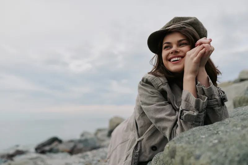 Smiling woman in warm hat and coat leans on rocks by the sea, looking away with a joyful expression. Overcast sky and calm water create a peaceful outdoor mood.