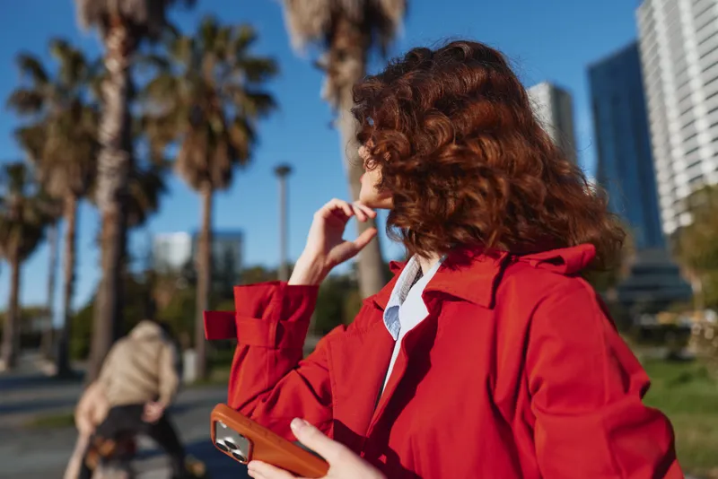 A woman with curly hair in a red coat stands outdoors in a sunny urban park, looking to the side with a thoughtful pose amid palm trees and modern buildings, fashion forward and confident.