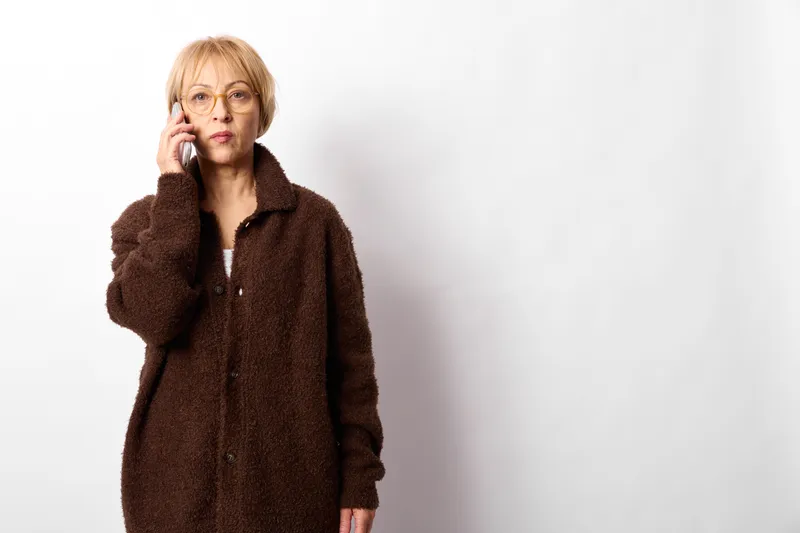 Elderly woman in brown coat, holding phone to ear, wearing glasses, studio portrait on white background, thoughtful expression, copy space, communication moment