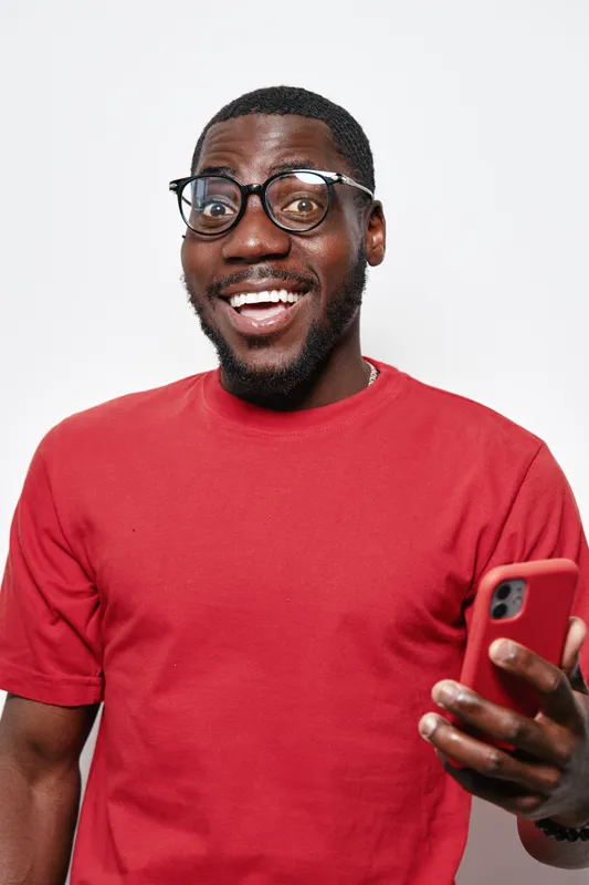 African American man with dark skin wearing glasses and red t shirt smiling and holding smartphone for communication and technology on white background.