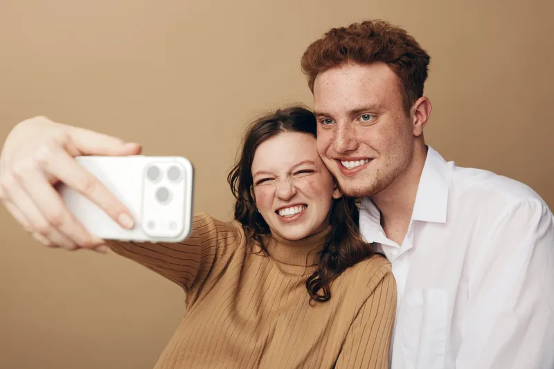 Young couple with red hair and brown hair takes a selfie together smiling. Casual clothing and beige background create a warm and happy mood for friendship and love concept.