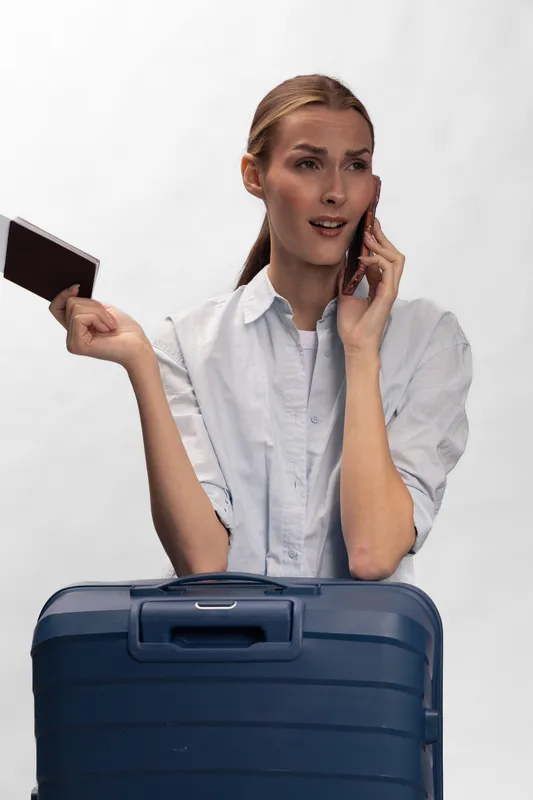 Young woman holding passport talks on phone with worried expression standing behind blue suitcase. Concept for travel issues, delayed flights, and travel planning problems.