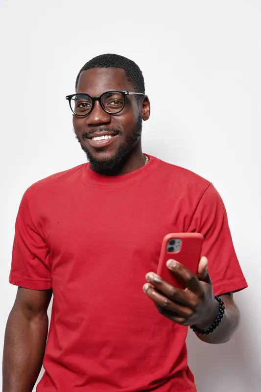 African American man with dark skin wearing glasses and red t shirt smiling while holding smartphone in hand isolated on white background. Casual style and technology use.