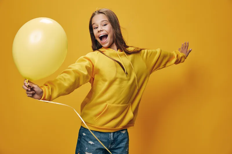 Girl, balloon, joy and smile with yellow hoodie standing on yellow background. Child, happiness and casual clothing with playful gesture for fun and celebration in studio.