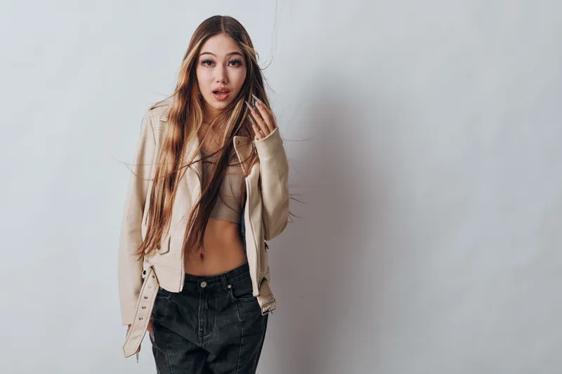 Young woman with long hair wearing light jacket and crop top standing against gray background looking at camera with casual confident expression in studio space.