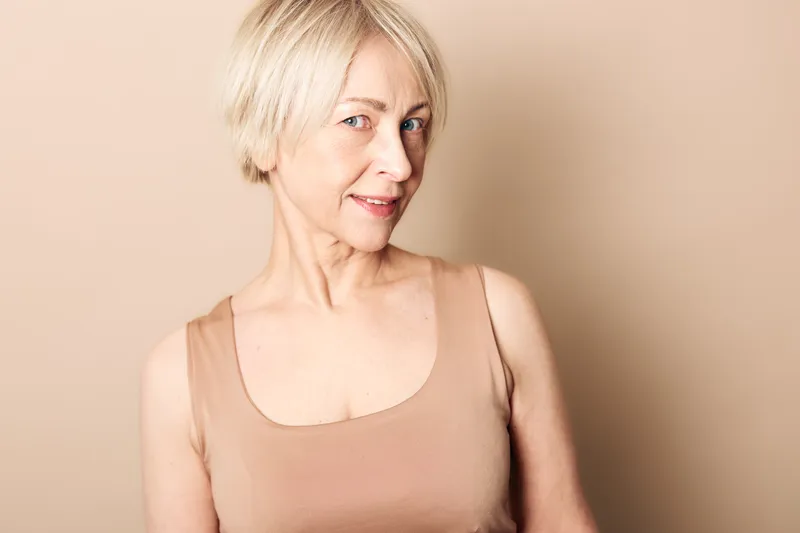 Confident mature woman with short blonde hair wearing a beige tank top smiles softly against a plain beige background. Concept for aging, beauty, and natural lifestyle.
