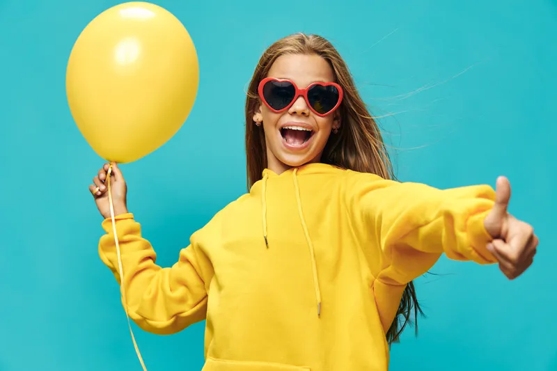 Girl in yellow hoodie and heart shaped sunglasses holding yellow balloon smiling and showing thumb up with blue background. Happy child, fun and cheerful mood portrait.