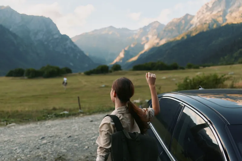 Woman traveler standing by car on mountain road, raising hand, scenic nature landscape, casual outfit, adventure concept, outdoor lifestyle.