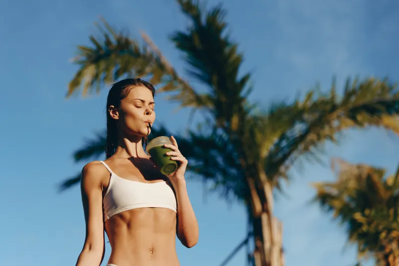 Young woman in a white crop top sipping a green smoothie outdoors under palm trees. Healthy lifestyle moment with hydration, wellness, and self care in sunny tropical light.